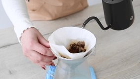 Close-up view of woman pouring hot water from black kettle on ground coffee lying on white filter in glass pitcher. Woman wears white sweater and beige apron. Slow motion video. Filter coffee theme.