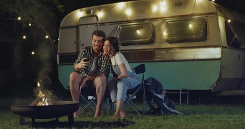 Cinematic shot of carefree couple in love having fun to make video call together to friends with smartphone near their trailer with lighting while enjoying romantic trip with camping caravan at night.