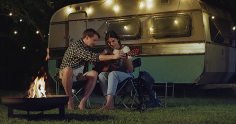 Cinematic shot of happy carefree husband is teaching to play guitar to his wife both sitting near their trailer with lighting while enjoying together romantic trip with camping caravan at night.