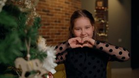 Cute child girl show heart by hands stay at christmas tree in living room at home. Young female person looking at camera in new year decorations indoors