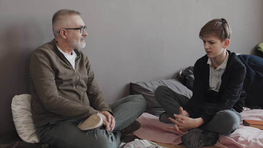 Full shot of senior Caucasian man and his grandson sitting on mattresses on floor of refuge, talking at daytime