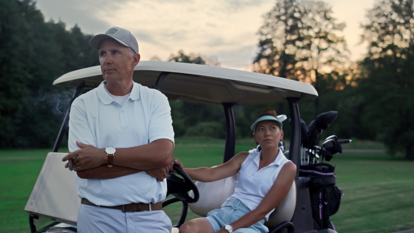 Wealthy golf players resting at sport cart. Sporty couple enjoy summer weekend close up. Thoughtful golfers group posing looking distance by golfing buggy equipment clubs. Luxury hobby leisure concept Royalty-Free Stock Footage #1090598573