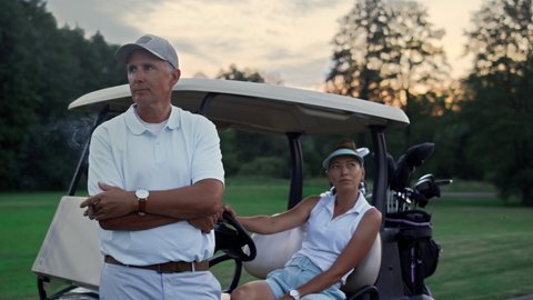 Wealthy golf players resting at sport cart. Sporty couple enjoy summer weekend close up. Thoughtful golfers group posing looking distance by golfing buggy equipment clubs. Luxury hobby leisure concept