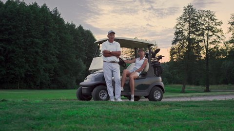 Two golfers rest outdoors on golf course. Married couple relax in golf cart. Wealthy sport team group posing look camera on fairway game. Professional players enjoy summer sunset. Weekend concept.