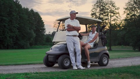 Couple posing golf cart outside. Two golfers take clubs sport equipment on sunset field. Wealthy married lovers enjoy relax activity on weekend. Golfing team in driver seat. Active leisure concept.