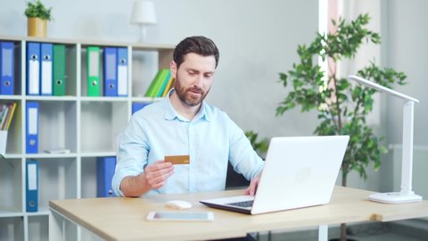 Happy cheerful smiling young adult man doing online shopping or e-shopping satisfied employee entrepreneur making online payment paying for service sitting with credit card at laptop in modern office