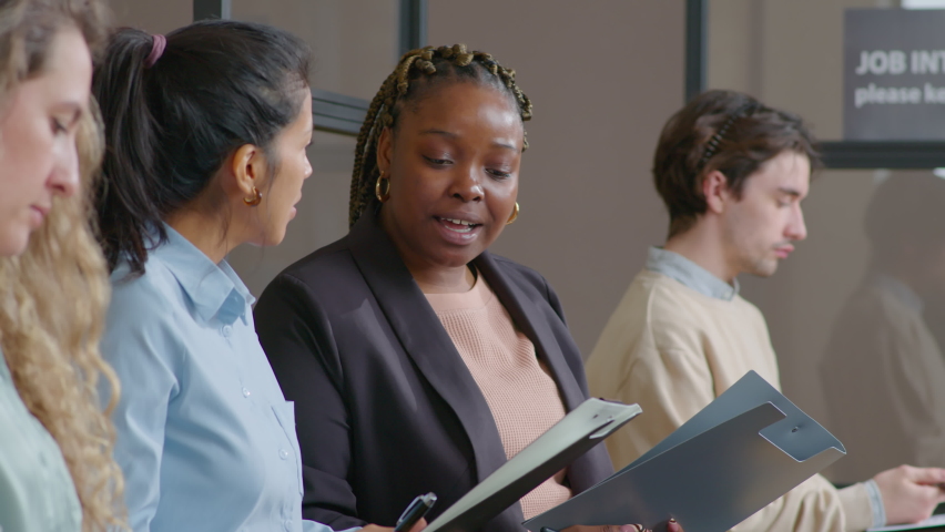 Tilt up shot of African American and Hispanic businesswomen discussing application form while sitting in line with other candidates and waiting for job interview
