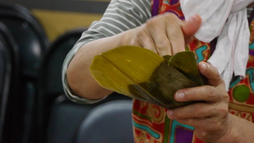 Hands of woman wrapping traditional purple rice triangle into leaf container during food prep, filmed in close up