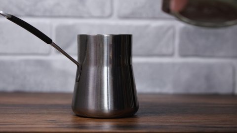 A man's hand with a wooden spoon pours fresh ground coffee into a steel cezve. In the background is a bright stone wall. The concept of Turkish coffee making