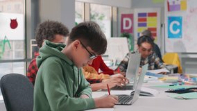 Teenage male student sit down at desk and write task in notebook using laptop with multiethnic classmates study on background - Powered by Shutterstock - Get 15% off with code: PIKWIZARD15