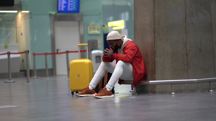 Sad African American man upset at airport his flight is delayed. Depressed traveler male waiting for a plane sitting in empty terminal with baggage. Exhausted guy on a long night connection at airport | Shutterstock HD Video #1090617119