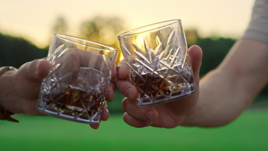 Friends toast glasses outdoors. Two men clinking drink together on golden sunset nature close up. Active unknown people celebrate cheering on grass field country club closeup. Luxury evening concept