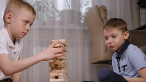 Close-up of two emotional school-age boys playing an exciting wooden blocks board game. The boy takes out a wooden block and the tower starts to wobble.