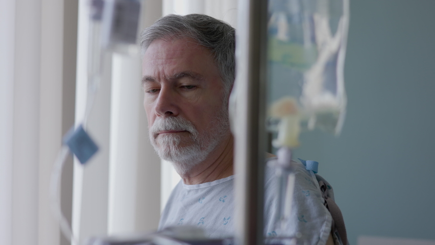 A solemn senior adult hooked up to an IV solution stands by his hospital room window framed between the plastic vacuum bags that are part of the intravenous delivery system. Royalty-Free Stock Footage #1090636241