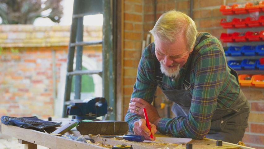 Senior man wearing overalls in sketching out design on workbench in home garage workshop - shot in slow motion Royalty-Free Stock Footage #1090641547