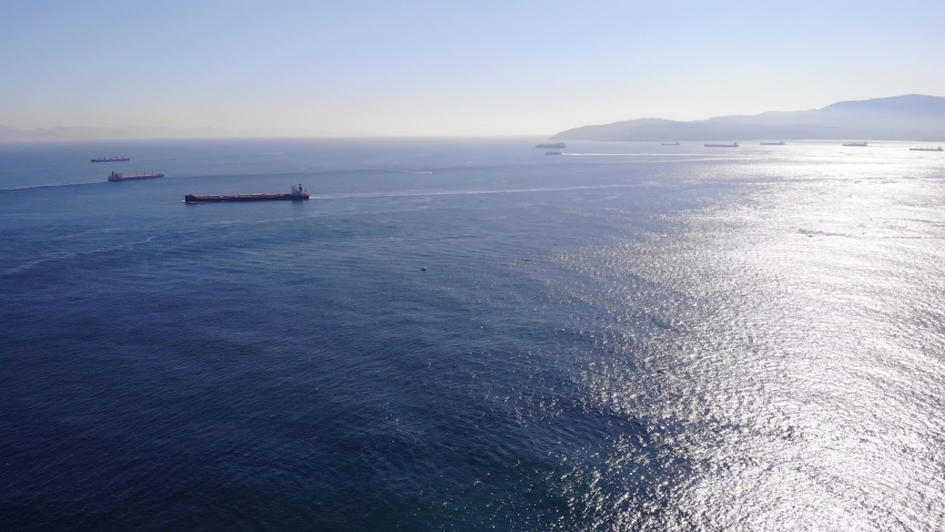 Large bulk carrier passes another freighter in the Strait of Gibraltar as several ships are anchored for Algeciras and a ferry in the background is moving at high speed to Africa. Drone panning shot