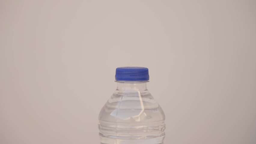 Closeup view 4k stock video footage of 500 ml transparent plastic bottle full of fresh water closed with blue plastic cap isolated on grey wall background. Woman trying to open bottle but cannot do it