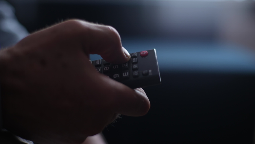 Close-up unrecognizable mature adult male hands holding TV remote control and switches TV channel. Closeup cropped shot of senior man flipping through TV channels with remote control, slow motion. Royalty-Free Stock Footage #1090651299