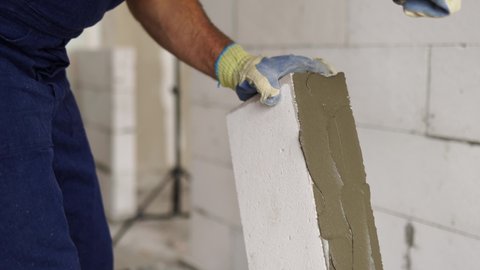 Bricklayer applies adhesive glue on autoclaved aerated concrete blocks with notched trowel. Brickwork worker contractor removes excess mortar from foam concrete doing masonry.
