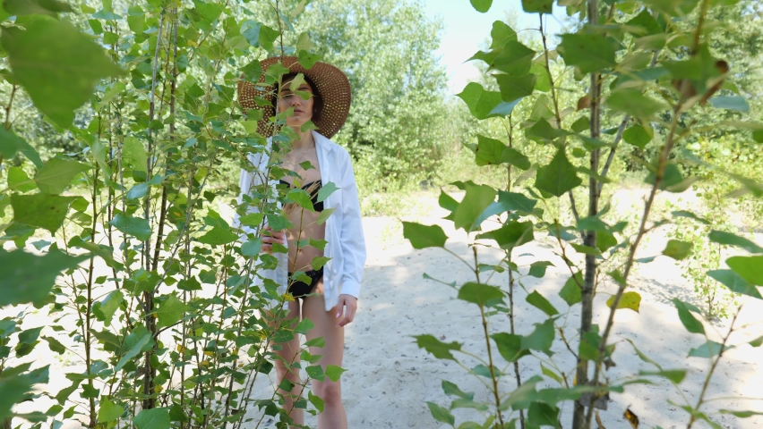 A young brunette woman in a black bathing suit and a beach hat walks on the sand near the green foliage. Woman in a white shirt on the sand