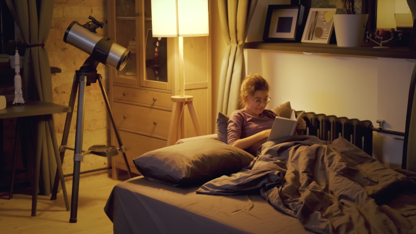 Caucasian girl wearing eyeglasses relaxing in bed in evening reading book about solar system before going to sleep, handheld shot