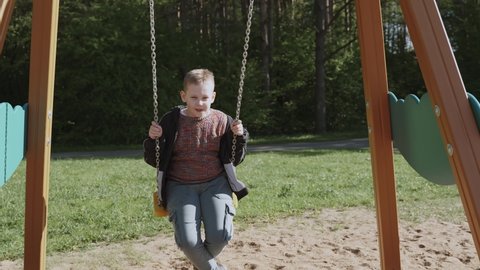 Caucasian school-age boy in windbreaker and jeans swings relaxed and calmly on wooden swing near the park, slow motion. Swings on chains and wooden supports on the sand.