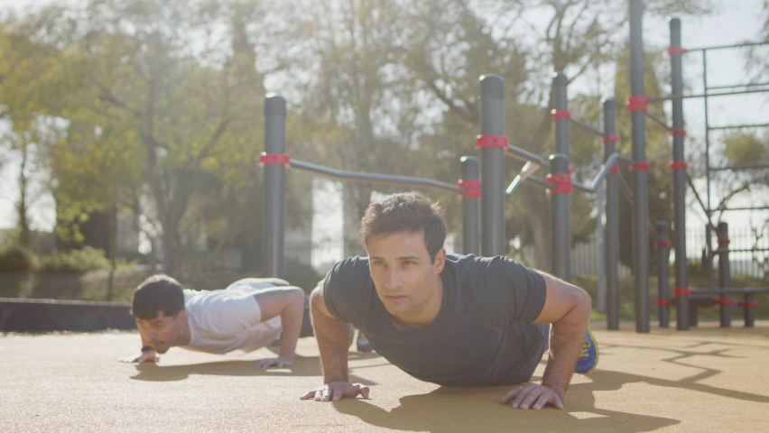 Concentrated men doing press ups on sports ground. Buddies training together in summer morning, motivating each other, devoting time to physical activity. Teamwork, outdoor workout, sport concept | Shutterstock HD Video #1090673451