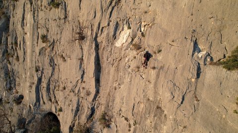 AERIAL: Rock climber equipped with rope ascending up the sunlit limestone wall. Man climbing up the wall and searching for good grips. Adrenaline outdoor activity in beautiful natural environment.