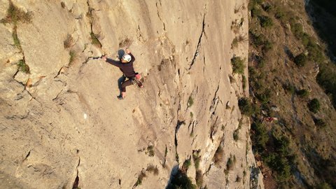 AERIAL: Young male rock climber with rope ascending up the sunlit limestone wall. Man climbing up the wall and searching for good grips. Adrenaline outdoor activity in beautiful natural environment.