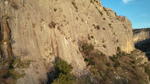 AERIAL: Flying close to sunlit rock climbing wall and approaching male climber. Adrenaline outdoor activity in beautiful and picturesque environment. Rock climber saluting in the middle of the wall.