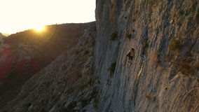 AERIAL: Woman climbing in the middle of the limestone wall with golden sun flare in background. Female climber ascending during sunset. Adrenaline outdoor activity in beautiful natural environment.