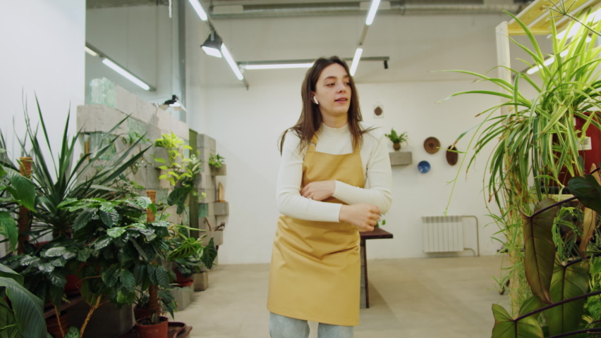 Florist woman dancing. Happy greenhouse worker listening to music, happy botanist having fun close-up, small business owner. Female gardener working in flower shop, plant store.  Royalty-Free Stock Footage #1090679485