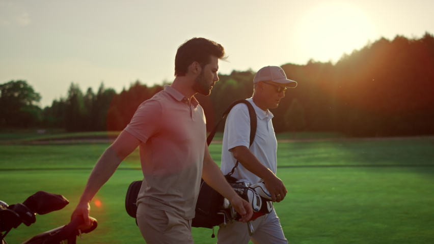 Businessmen walking golf course outside. Two professional players carry clubs in sportswear. Golfing team friends discuss wealthy lifestyle hobby on summer sunset field. Sport partnership concept. Royalty-Free Stock Footage #1090684051