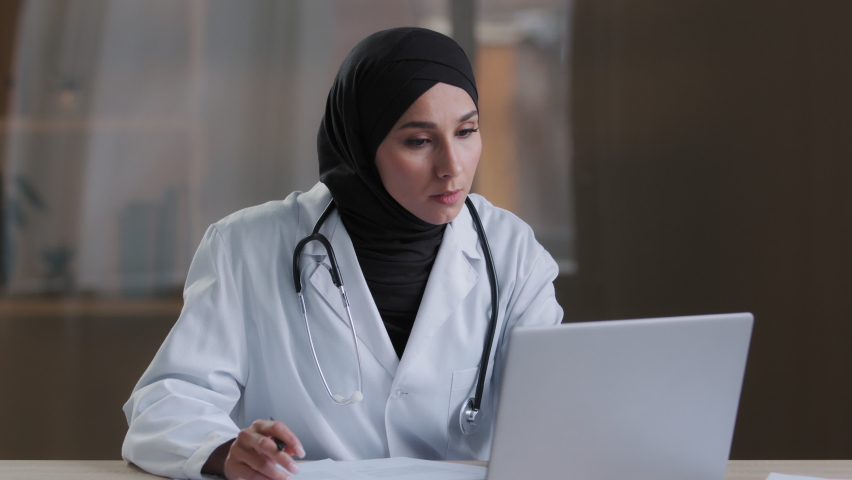 Concentrated muslim arabian female doctor chief physician practitioner signs documents sit at clinic cabinet search information on laptop use computer virtual medical service write recipe prescription Royalty-Free Stock Footage #1090685431