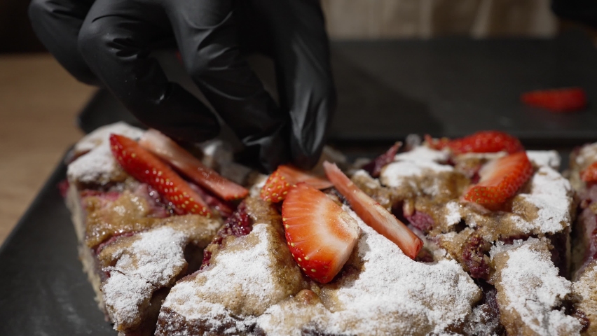 Pastry cook puts fresh strawberry slices on cake sprinkled with sugar powder. Woman decorates fresh baked cake with strawberry slices closeup