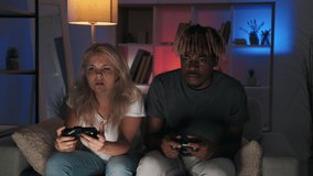 Night video game. Home fun. Late entertainment. Confused beginner couple enjoying learning playing using joystick controller on couch in dark living room interior with color light.