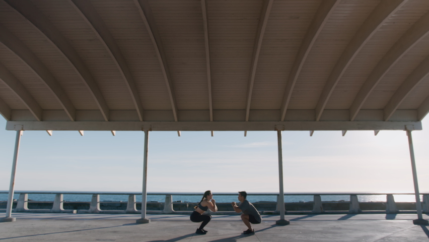 Shot of two people, man and woman, friends, coach working out together, making gym exercises on warm sunny daytime at outside park near sea or ocean water, beach at beautiful urban background place