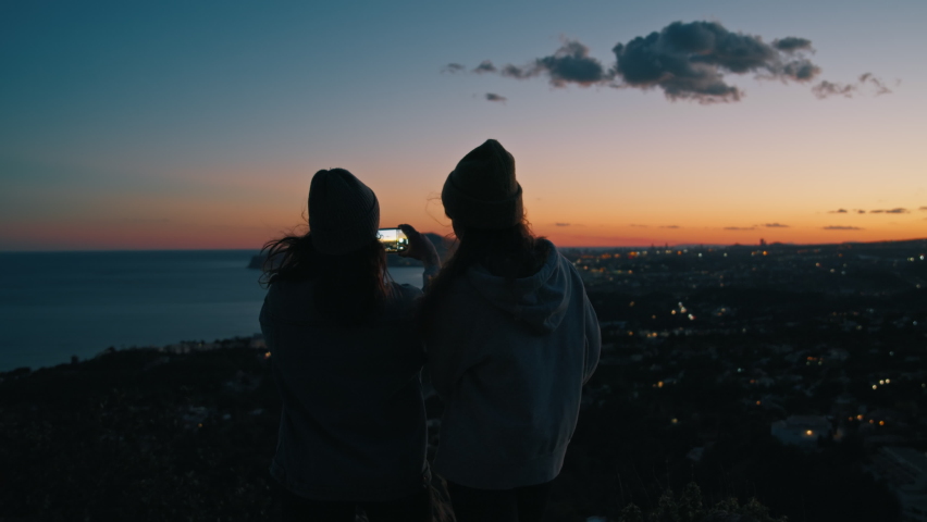 Young women, millennial girls, friends silhouette take colorful beautiful dark sunset seashore panoramic photo holding phone together, using camera on smartphone during weekend, trip, travel, vacation