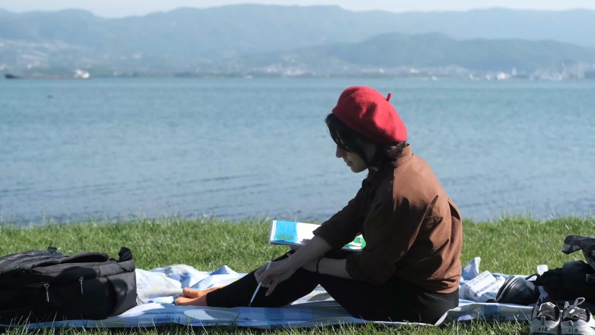 Youg women artist sitting against the sea view and painting, woman sitting on the grass by the seascape and painting on canvas
