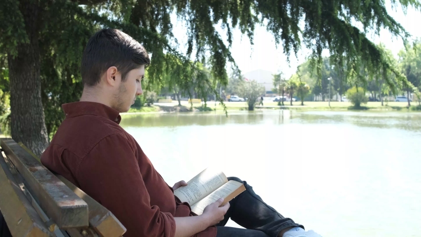 Young man sitting by the river reading a book, spending time on the bench in the shade under the trees, relaxing by reading book in the green area