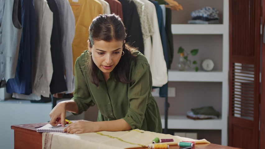 Modern creative Indian Asian woman or female professional fashion stylist is thinking and measuring cloth with tape busy working in her garments design boutique. Women empowerment and passion concept | Shutterstock HD Video #1090719871