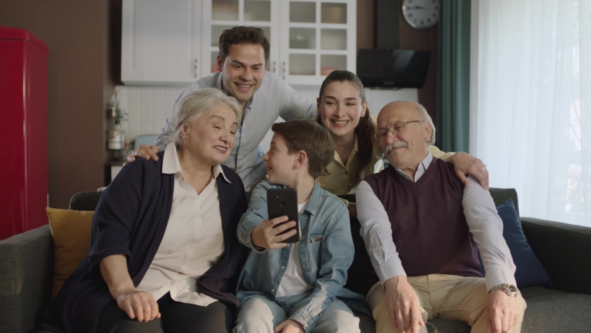 Young couple with children, their son and elderly parents sitting on sofa in living room, taking selfie together.Portrait of happy cheerful big family smiling at their smartphones in cozy living room.