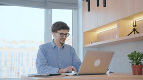 Young man in headphones and glasses using a laptop positively and with a smile communicates via video chat. A man works remotely with colleagues at work through a laptop using video communication