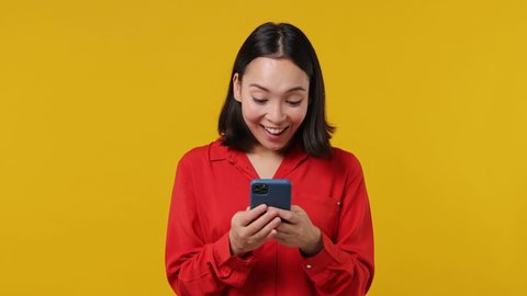 Amazed excited young woman of Asian ethnicity 20s wears red shirt hold in hand use point finger on mobile cell phone showing thumb up like gesture isolated on plain yellow background studio portrait