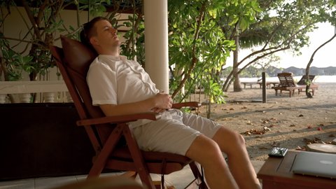 A man rests on a chair on the shores of the Indian Ocean, wearing glasses. The concept of a sleep break while working on a tropical island