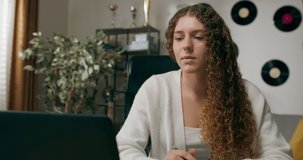 Girl with curly hair sits at desk in living room and studies on laptop taking notes focused young woman work on computer writes in notebook takes online course or training at home concept of education