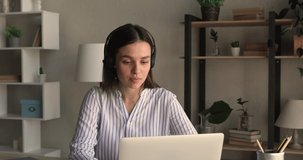 Young female employee sit at workplace desk looks at laptop screen talking to client distantly, wear headset provide professional technical support to customer remotely using video call application
