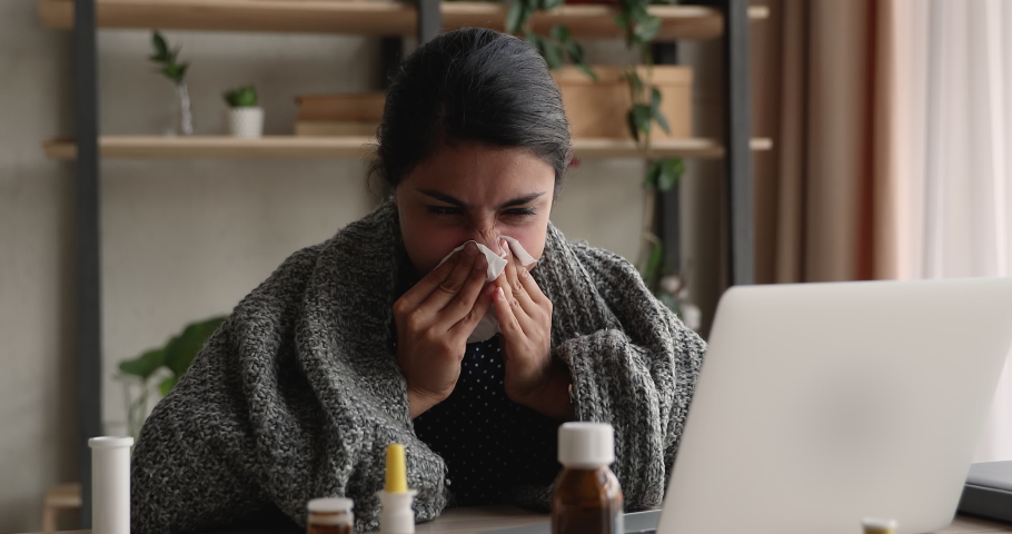 Indian businesswoman sit at workplace wrapped in warm plaid, blow runny nose in paper tissue, hard to breathe feels unhealthy look sick, meds on desk. Seasonal grippe, employee need sick leave concept | Shutterstock HD Video #1090734475