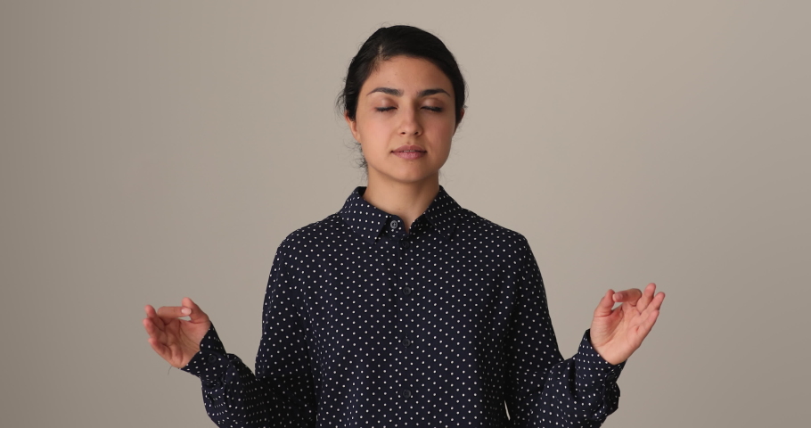 On gray studio background Indian ethnicity woman closed eyes folding fingers makes mudra gesture, do meditation practice, manage stress, increasing self-awareness, reducing negative emotions concept Royalty-Free Stock Footage #1090734491