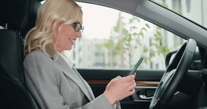 Smiling blond woman chatting or browse videos or photos or social networks on smartphone while sitting in car.